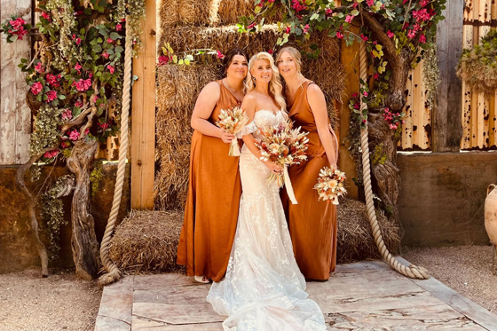 Bride and two bridesmaids wearing rust-coloured dresses and holding bouquets smile in front of hay bales